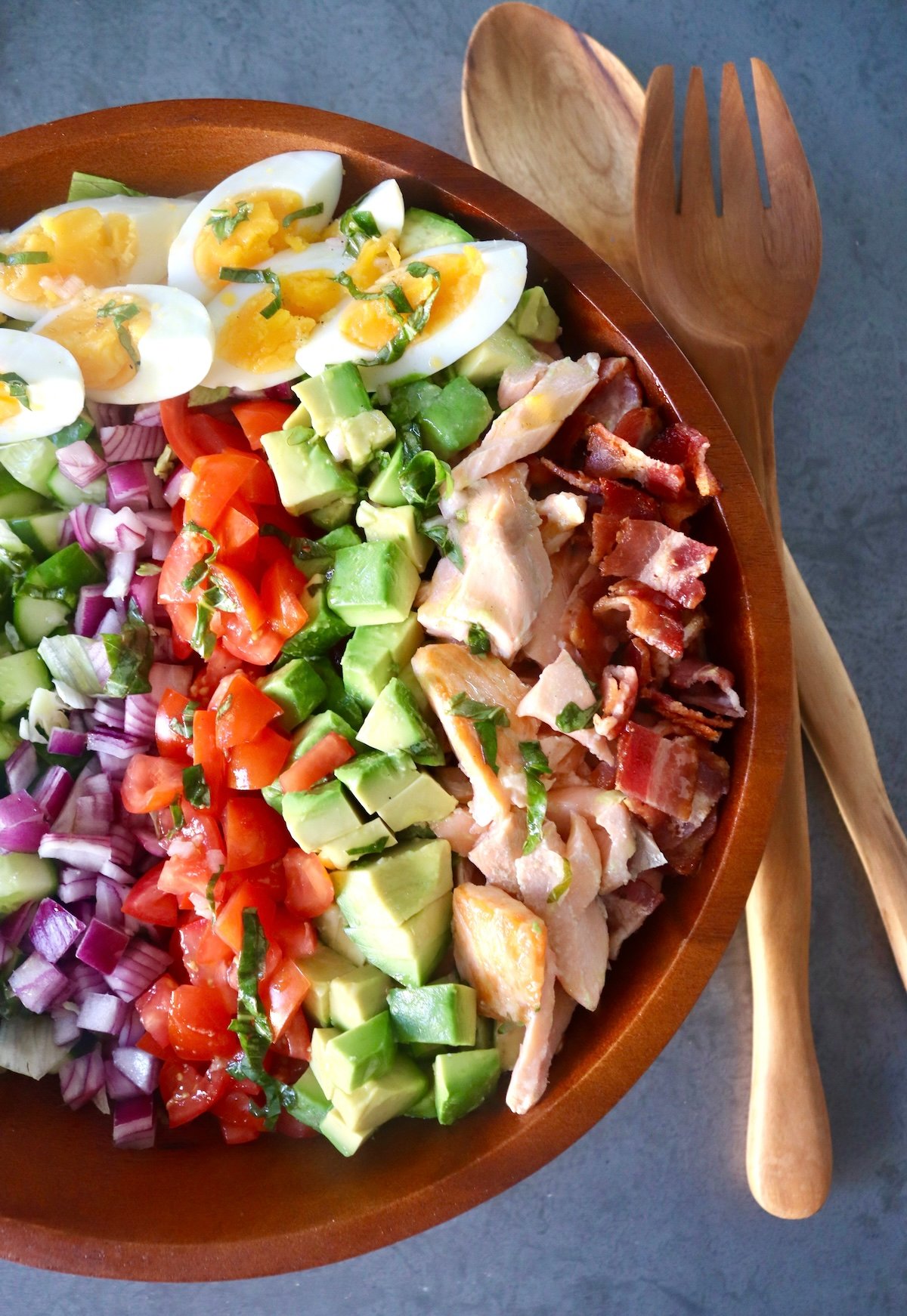 Salmon-Cobb-Salad Wooden bowl filled with a composed salmon cobb salad, with rows of tomato, salmon, cucumber, red onion, basil, salmon and hard boiled eggs, with wooden serving utensils next to it.
