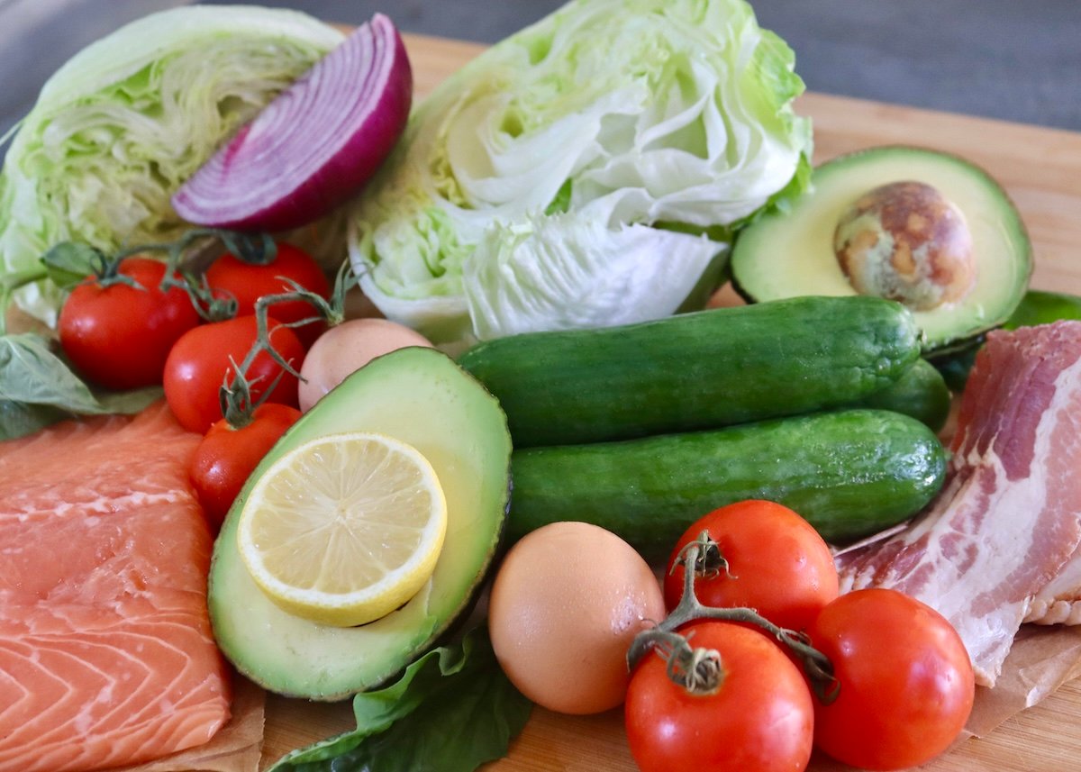Raw salmon, avocado, lemon, tomatoes, raw bacon, cucumbers, iceberg lettuce, red onion and eggs on a cutting board.