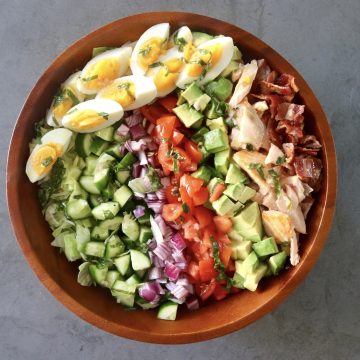 Top view of wooden bowl filled with a composed salmon cobb salad, with rows of tomato, salmon, cucumber, red onion, basil, salmon and hard boiled eggs.