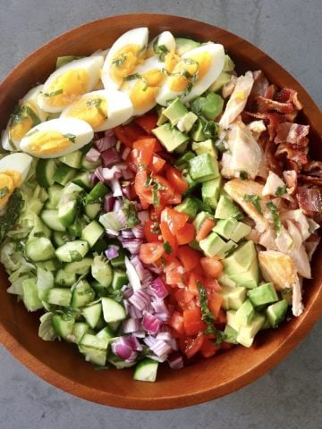 Top view of wooden bowl filled with a composed salmon cobb salad, with rows of tomato, salmon, cucumber, red onion, basil, salmon and hard boiled eggs.