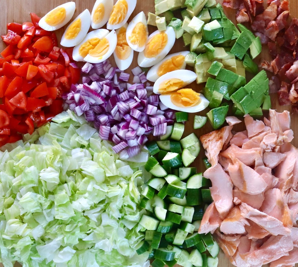 Cutting board full with chopped tomatoes, cucumbers, avocados, red onion, lettuce and quartered hard-boiled eggs.