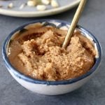 Small dark-blue-rimmed bowl with macadamia nut butter and a place behind it with the butter spread on toast, with a few sprinkled whole nuts.