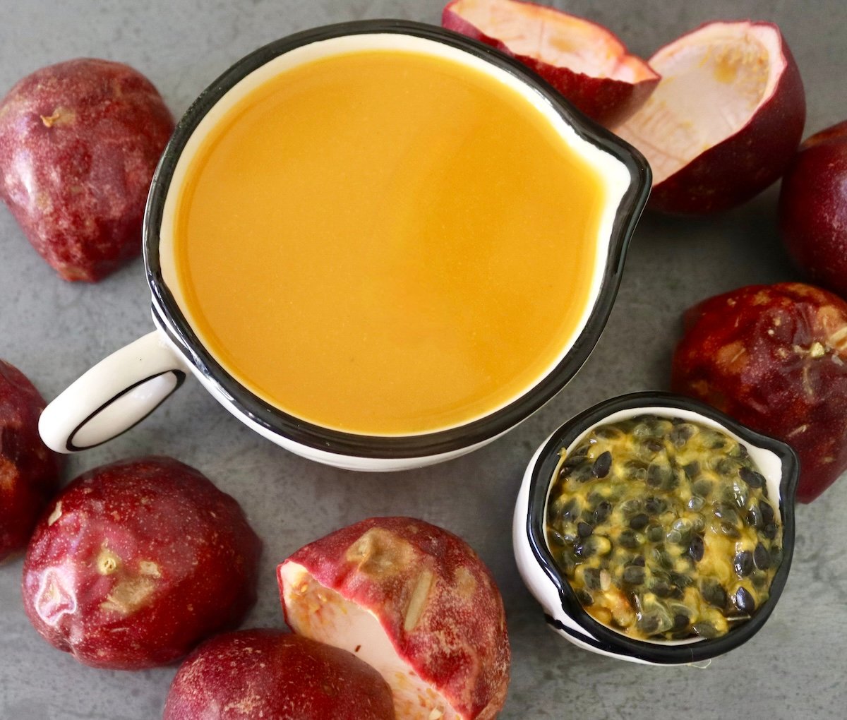 White mug with passion fruit juice, a smaller one with passion fruit seeds, and passion fruit skins all round them. 