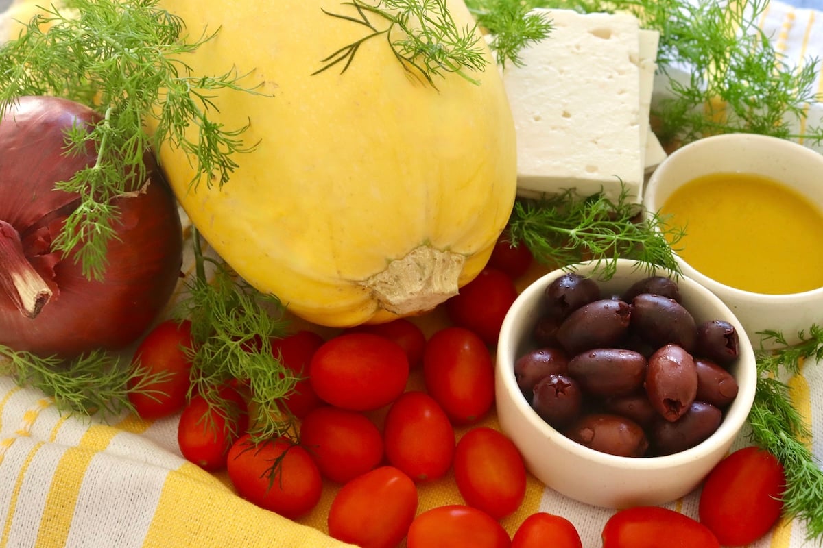 Spaghetti-Squash-Rings-with-Feta-and-Olives Whole spaghetti squash, teardrop tomatoes, Feta cheese block, red onion, olive oil, Kalamata olives and fresh dill all on a yellow and white cloth.
