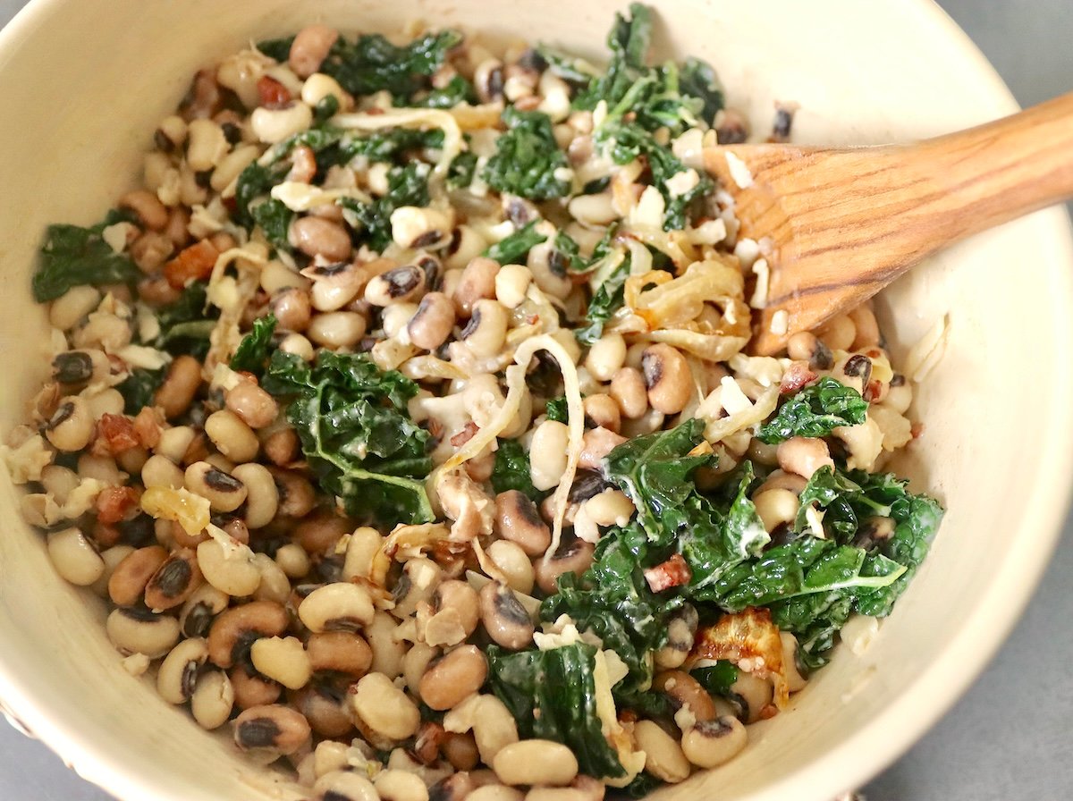 Black-eyed peas, kale, sliced onions, pancetta, and grated cheese in a mixing bowl with a wooden spoon.