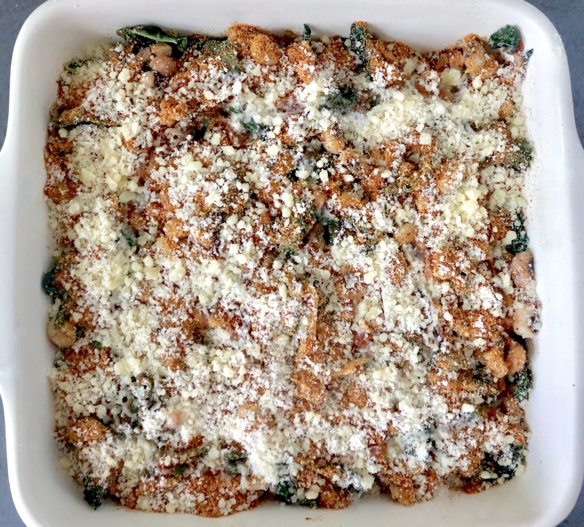 Black-eyed peas, kale, sliced onions, pancetta, and grated cheese in a square baking dish with breadcrumbs and grated cheese on top.