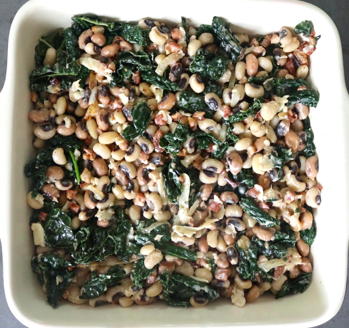 Black-eyed peas, kale, sliced onions, pancetta, and grated cheese in a square baking dish.