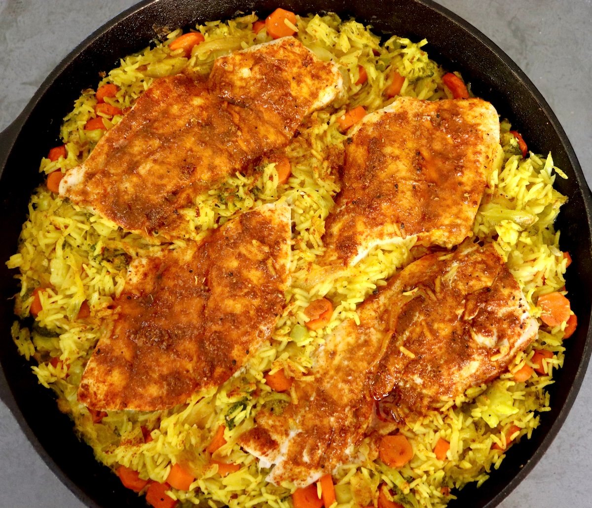 Baked-Red-Snapper-Recipe Cast iron skillet filled with yellow vegetable rice and 4 baked spice-coated fish fillets on top.