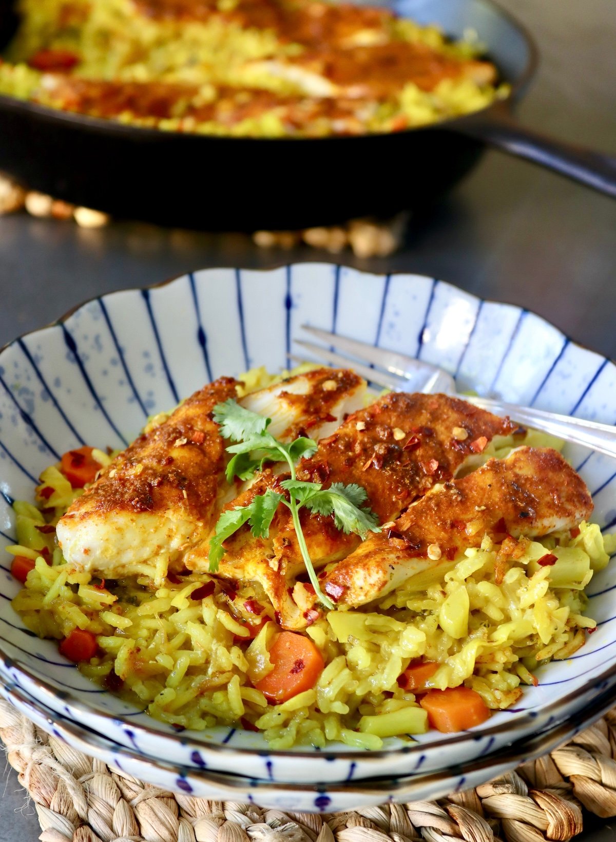 Blue striped plate with yellow rice and sliced, red-spiced fish, with a sprig of cilantro, and a cast iron skillet behind it.