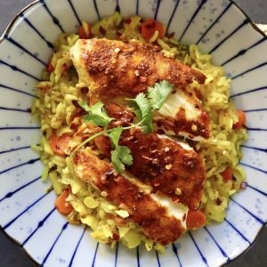 Top View of blue striped plate with yellow rice and sliced, red-spiced fish, with a sprig of cilantro.