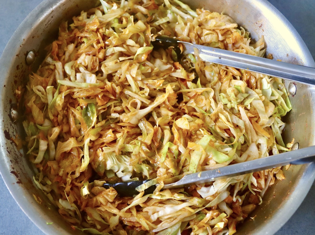Sautéed and wilted cabbage with tomato sauce in a sauté pan with kitchen tongs.