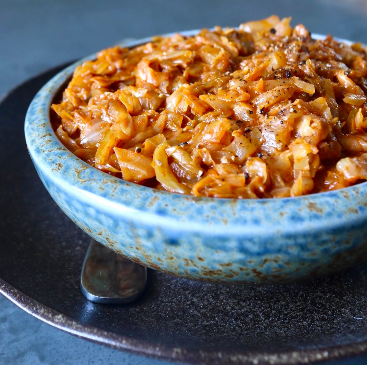 Light blue ceramic bowl full of tomato sautéed cabbage, on a dark brown ceramic plate.