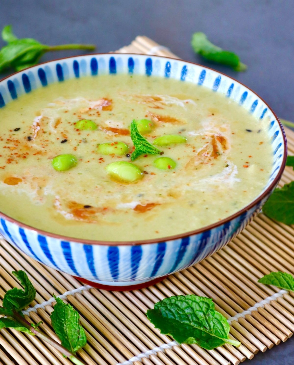 Edamame-Soup-Recipe Blue and white striped, ceramic bowl with edamame soup with chili oil, mint leaf, and a bit of cream.