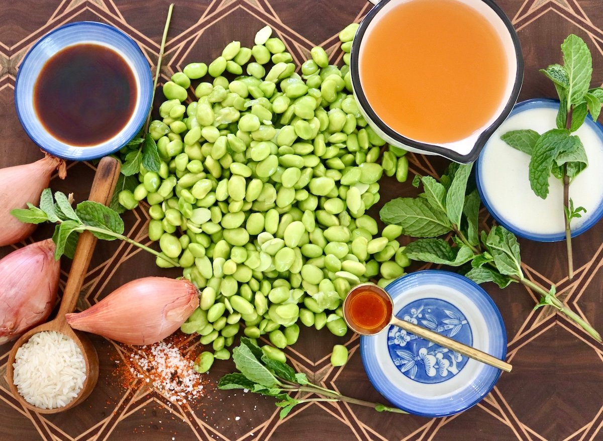 Edamame-Soup-Recipe Dark wood patterned cutting board with pile of edamame beans, 3 shallots, and bowls of soy sauce, cream, vegetable stock, mirin, and fresh mint.