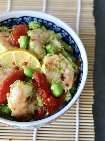 Ceramic bowl with colorful flowers on a sushi mat, filled with shrimp, tomatoes and edamame over rice.