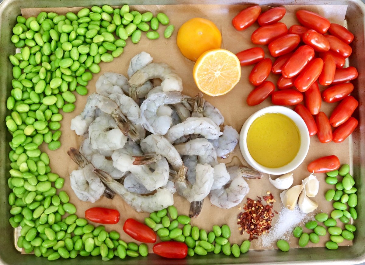Sheet-Pan-Shrimp-with-Edamame-Pesto Parchment paper-lined sheet pan with fresh edamame (shelled), raw shrimp, small tomatoes, bowl of olive oil, garlic cloves, crushed red pepper and lemon - all laid out separately.