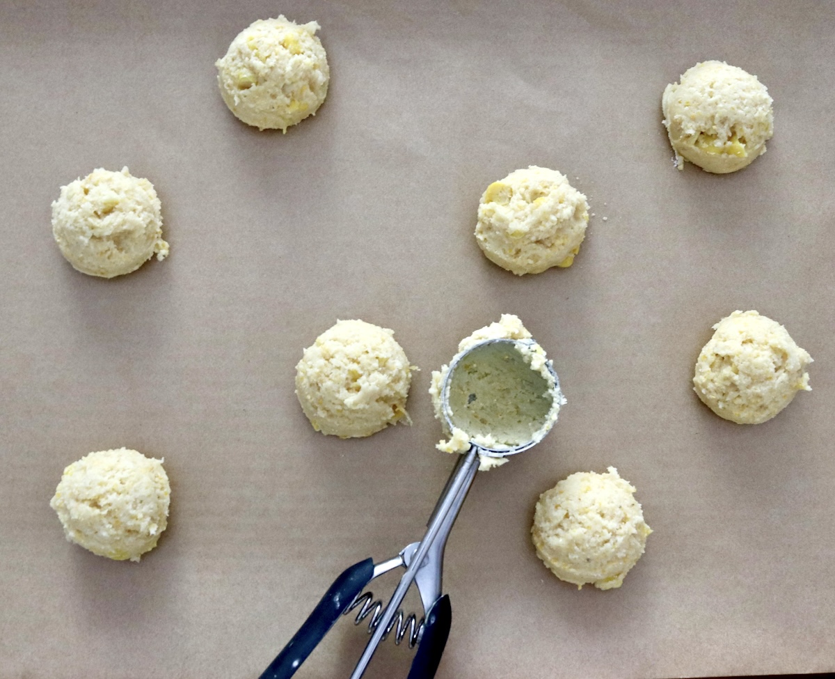 Several balls of cornbread cookie batter on a parchment-lined sheet pan with a cookie scoop.
