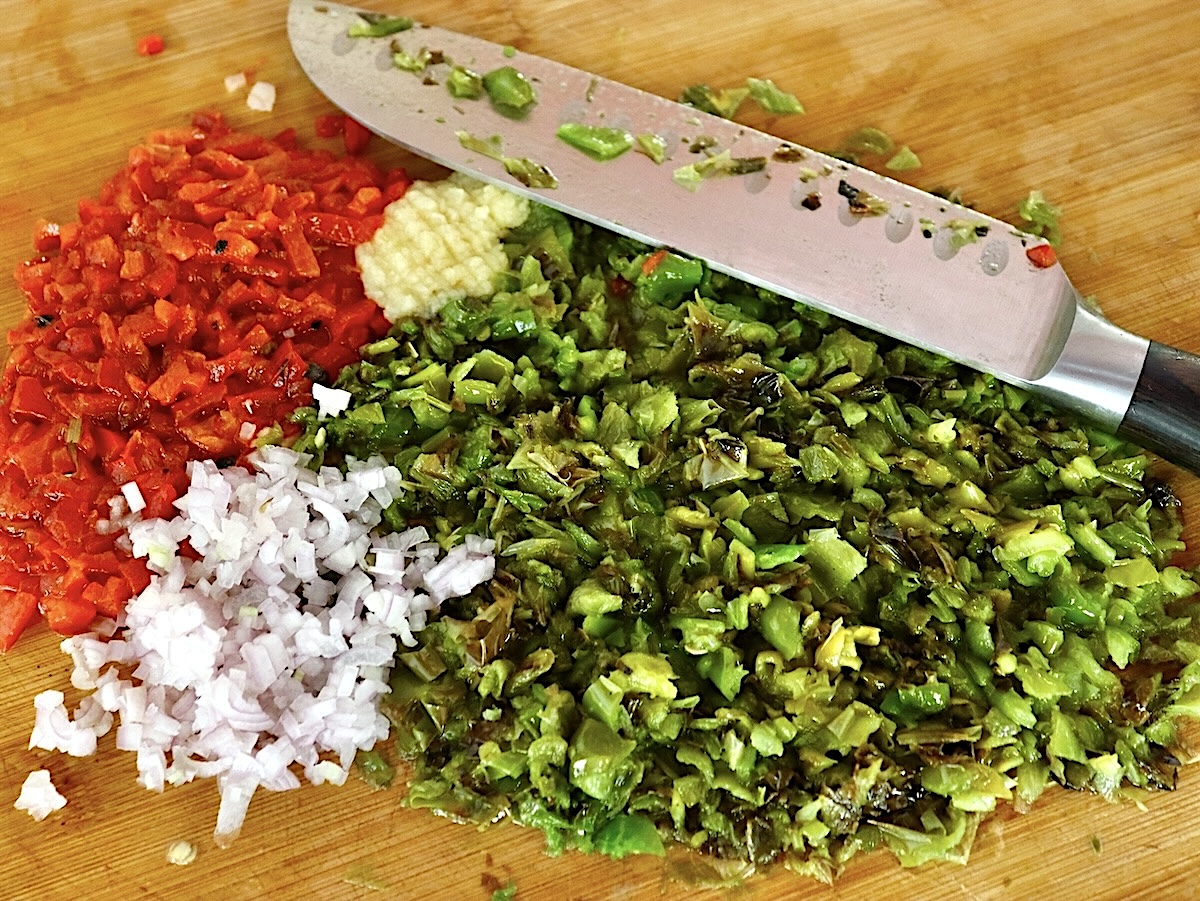 Shishito-Pepper-Salsa-Recipe Finely chopped red pepper, green shishito pepper, shallots and minced garlic on a cutting board with a chef's knife.