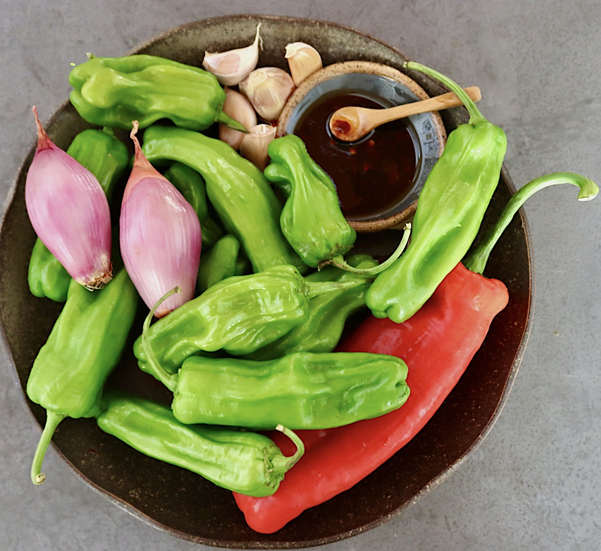 Shishito-Pepper-Salsa-Recipe Black plate with raw shishito peppers, one long red pepper, shallots and garlic cloves, with a tiny bowl of soy sauce.