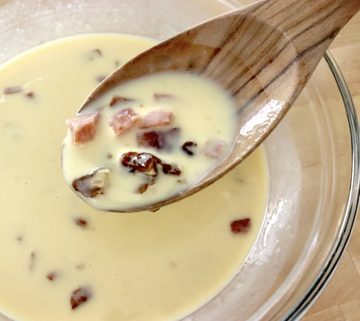 Glass bowl with whisked egg-milk mixture that includes diced dates and Spam, with a wooden spoon holding some over it.