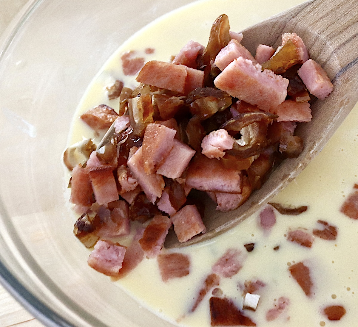Wooden spoon full with diced Spam and dates, held above glass bowl with whisked egg-milk mixture.