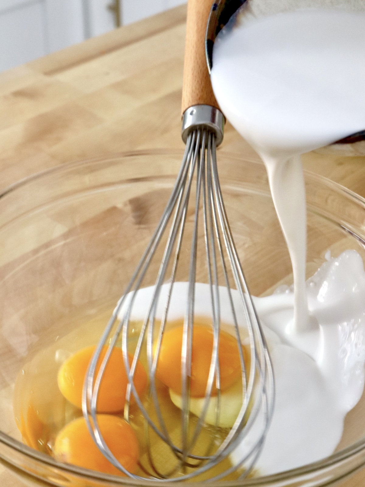 Glass bowl with a few eggs, and coconut milk being poured into it.