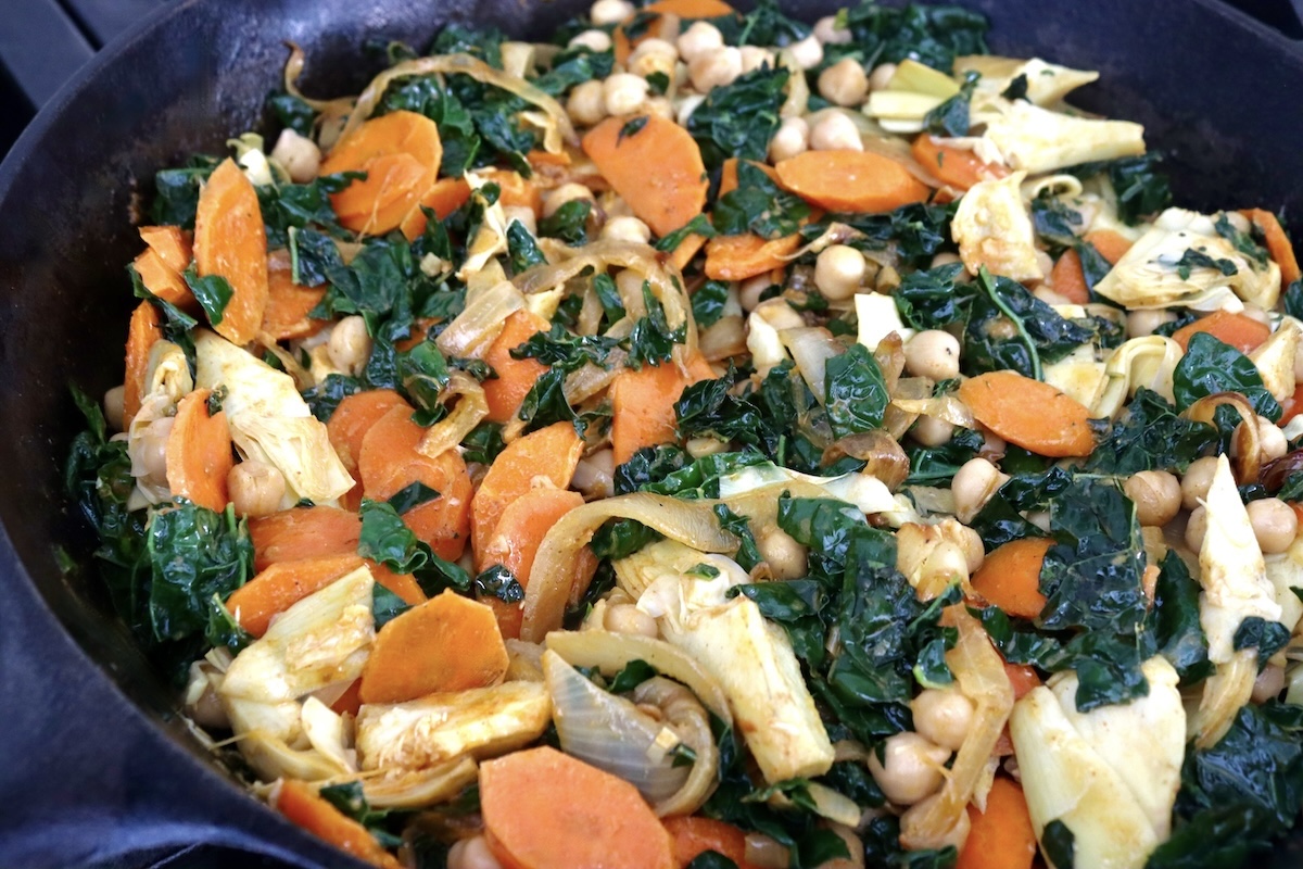 Whole-Cauliflower-One-Pan-Dinner-with-Tahini-Sauce Sautéed, sliced onions, carrots, kale and artichoke hearts in a cast iron skillet.