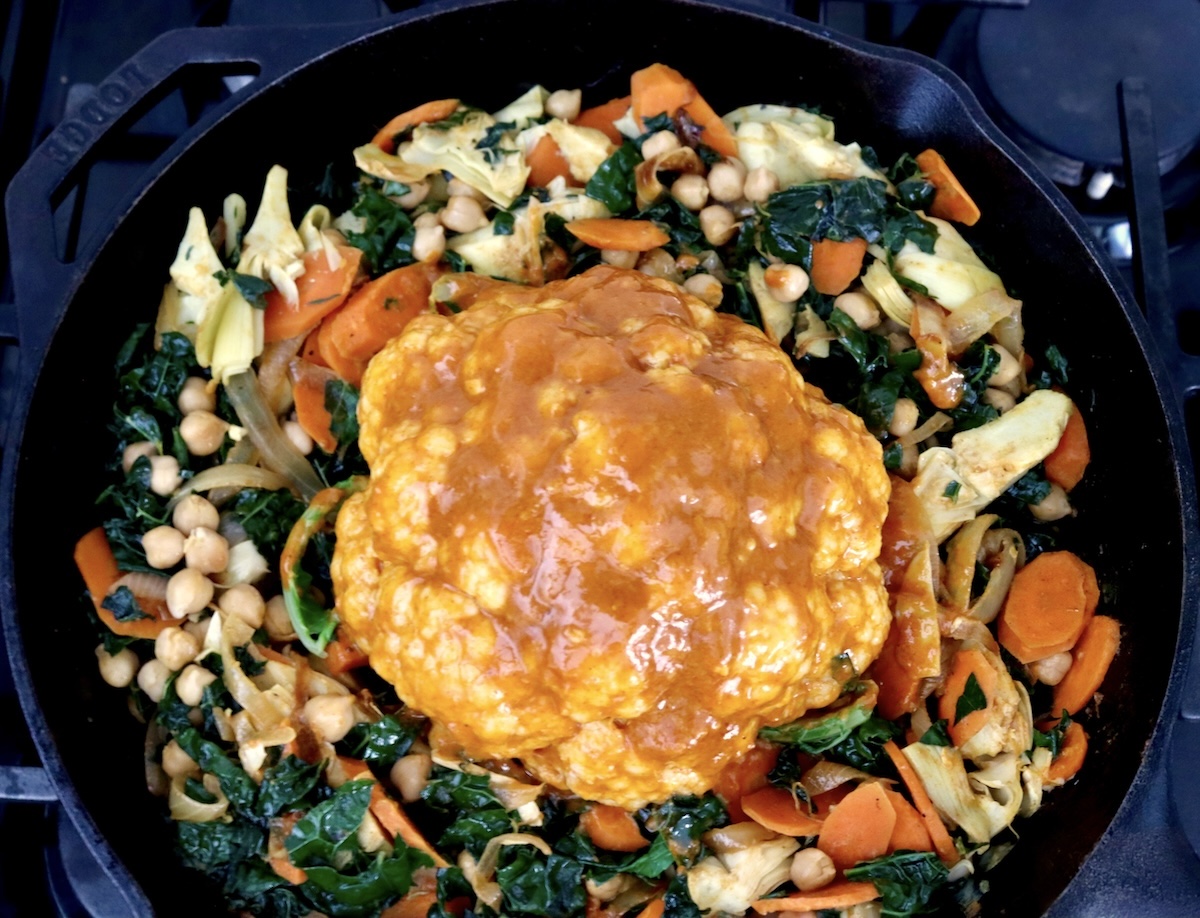 Whole-Cauliflower-One-Pan-Dinner-with-Tahini-Sauce Cast iron skillet with a ring of mixed vegetables around a whole head of cauliflower covered with red tahini sauce.