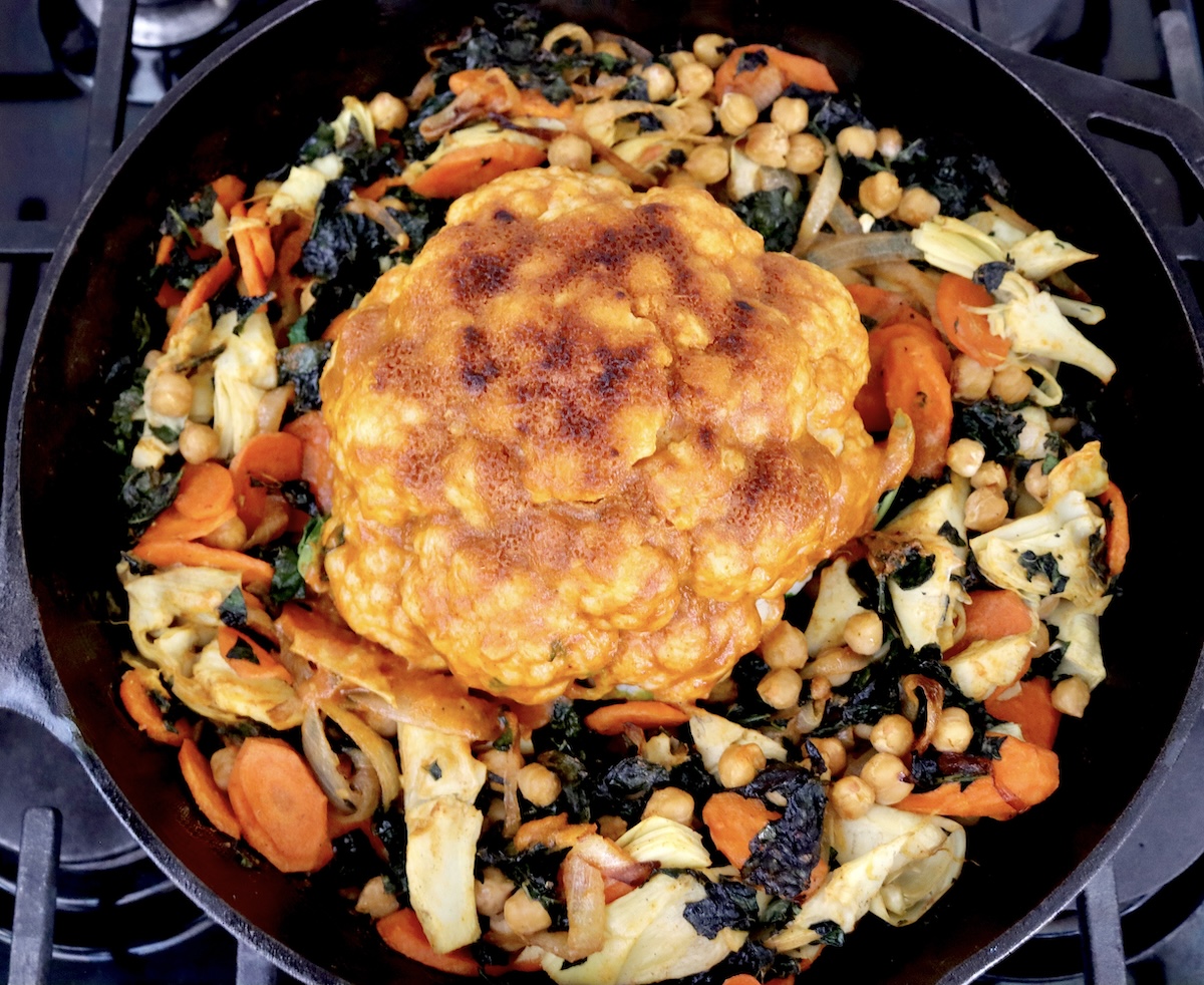 Whole-Cauliflower-One-Pan-Dinner-with-Tahini-Sauce Cast iron skillet with a ring of mixed vegetables around a broiled whole head of cauliflower covered with red tahini sauce.
