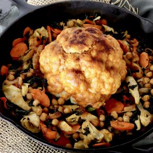 Whole cauliflower with red sauce in the center of a cast iron skillet with other vegetables.