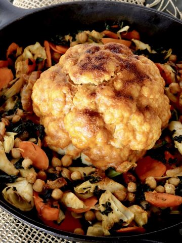 Whole cauliflower with red sauce in the center of a cast iron skillet with other vegetables.