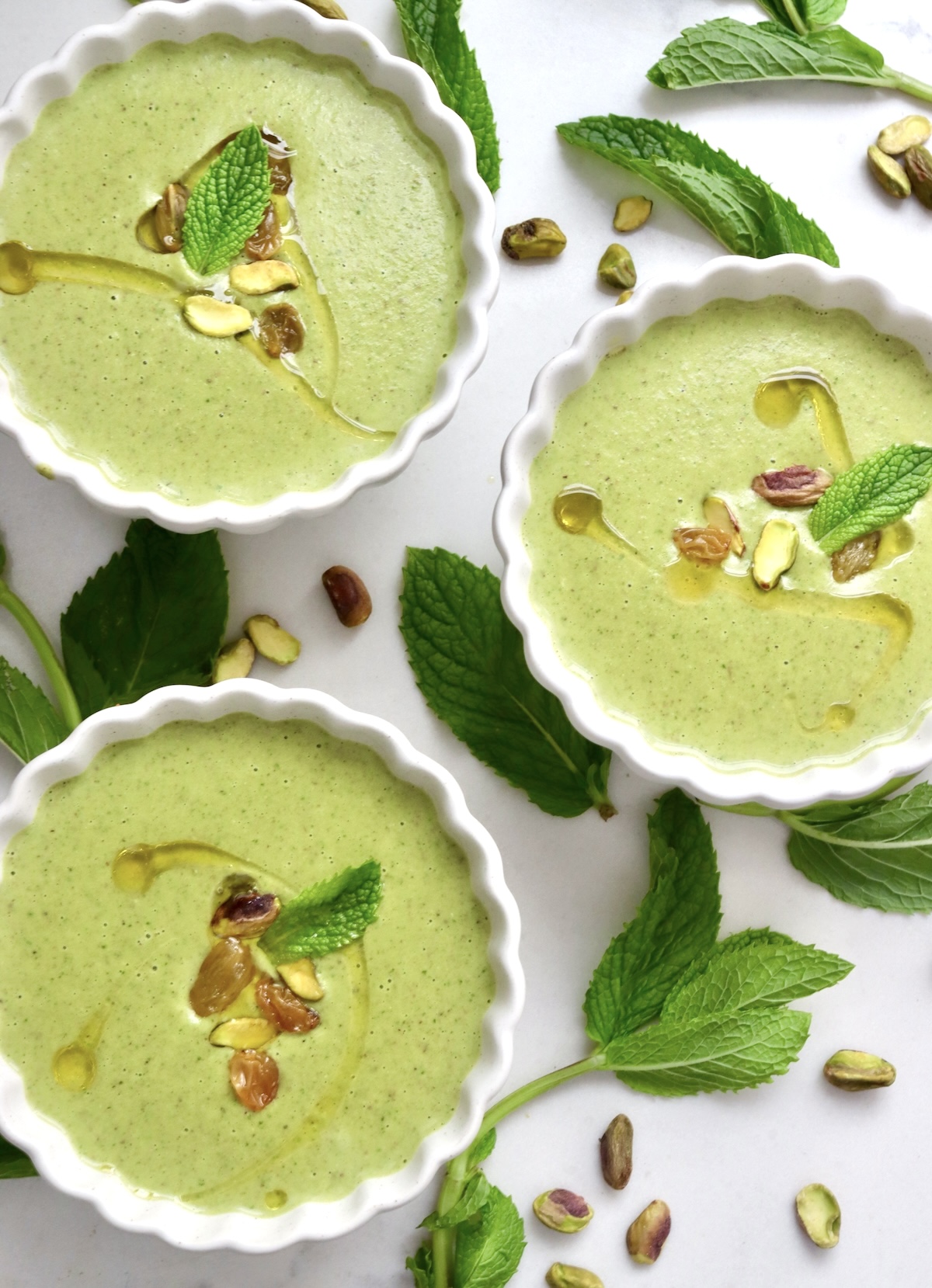 Top view of three white crimp-edged bowls with minty green pistachio soup with a few garnishes including mint leaves.