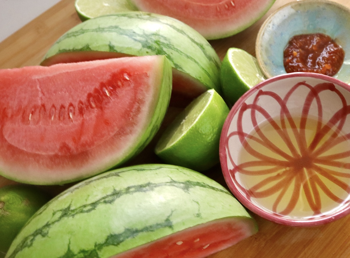 Lime-Chile-Grilled-Watermelon Mini watermelon quartered with pink and white painted ceramic bowl with oil, small blue ceramic bowl with chili paste and a couple of limes cut in half.