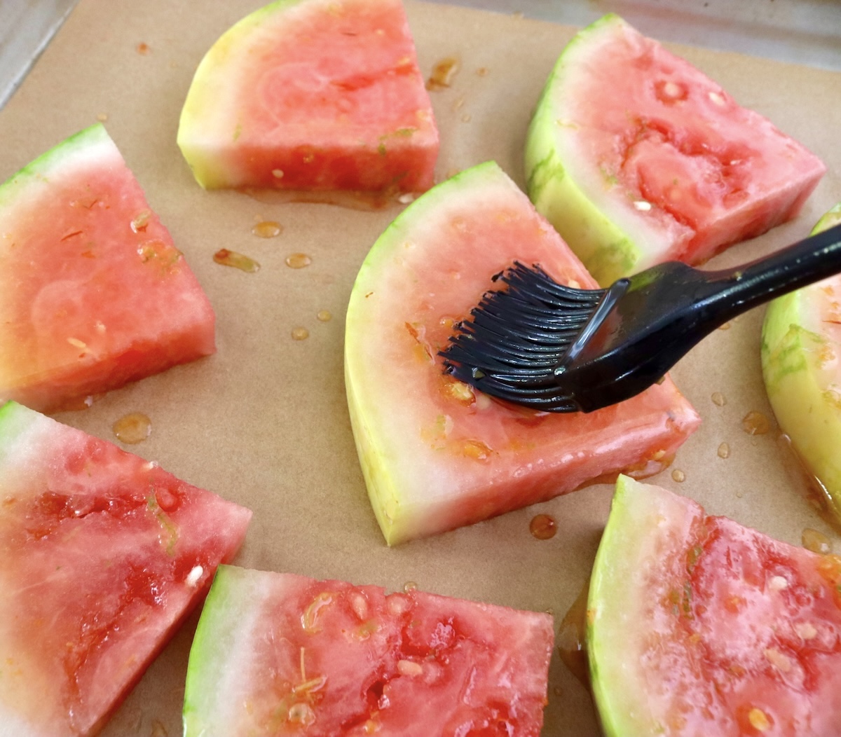 Lime-Chile-Grilled-Watermelon 1-inch slices of watermelon on parchment paper with a marinated of lime and chile being added with a black pastry brush.