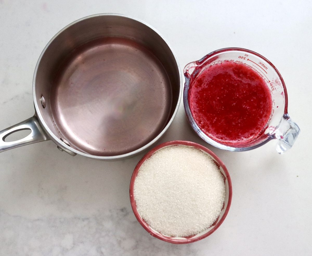 Prickly-Pear-Syrup Small saucepan with water, red-rimmed ceramic bowl with granulated sugar and glass measuring cup with prickly pear juice, on a white marble countertop.